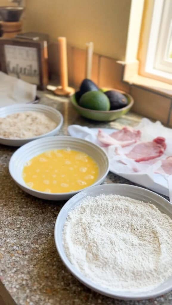 Dredging station set up for the Crispy Pork Cutlets for Pork Milanese featuring flour, eggs, and breadcrumbs in shallow bowls.