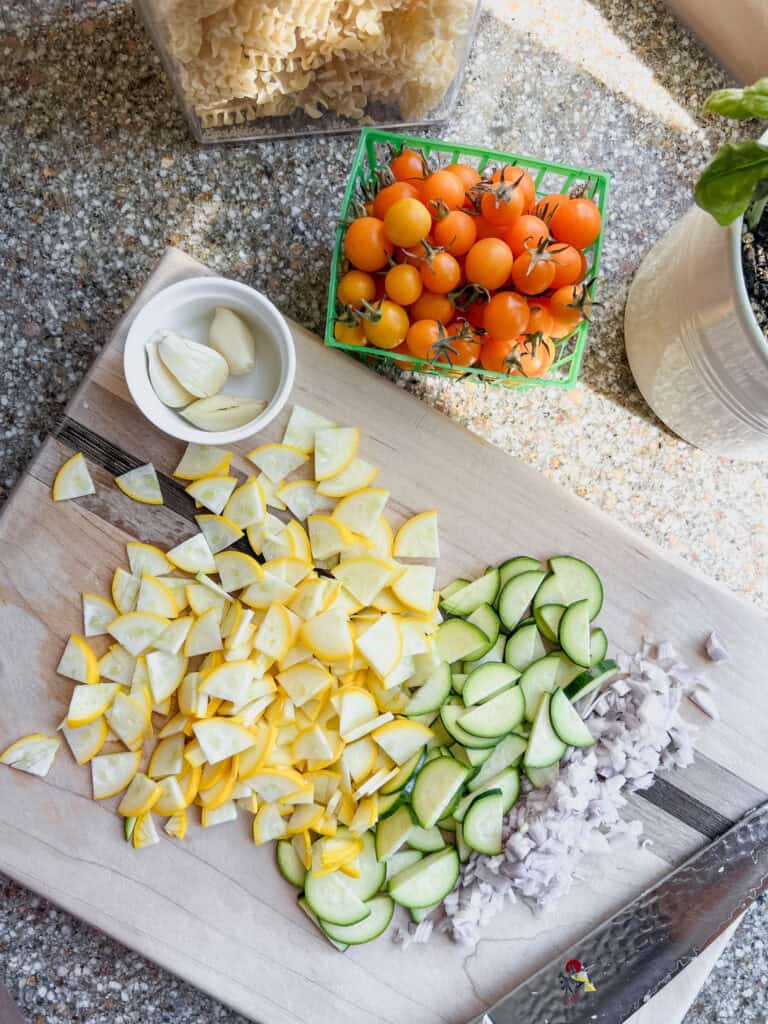 Ingredients for the sun gold tomato pasta recipe featuring sun gold tomatoes in a green basket, thinly sliced zucchini, finely chopped shallots, minced garlic, and a bowl of dried pasta. 