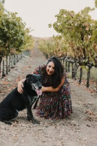 Photo of Frances in a floral dress kneeling beside her black labrador for an autumn photo against a backdrop of a vineyard with leaves of green and gold.