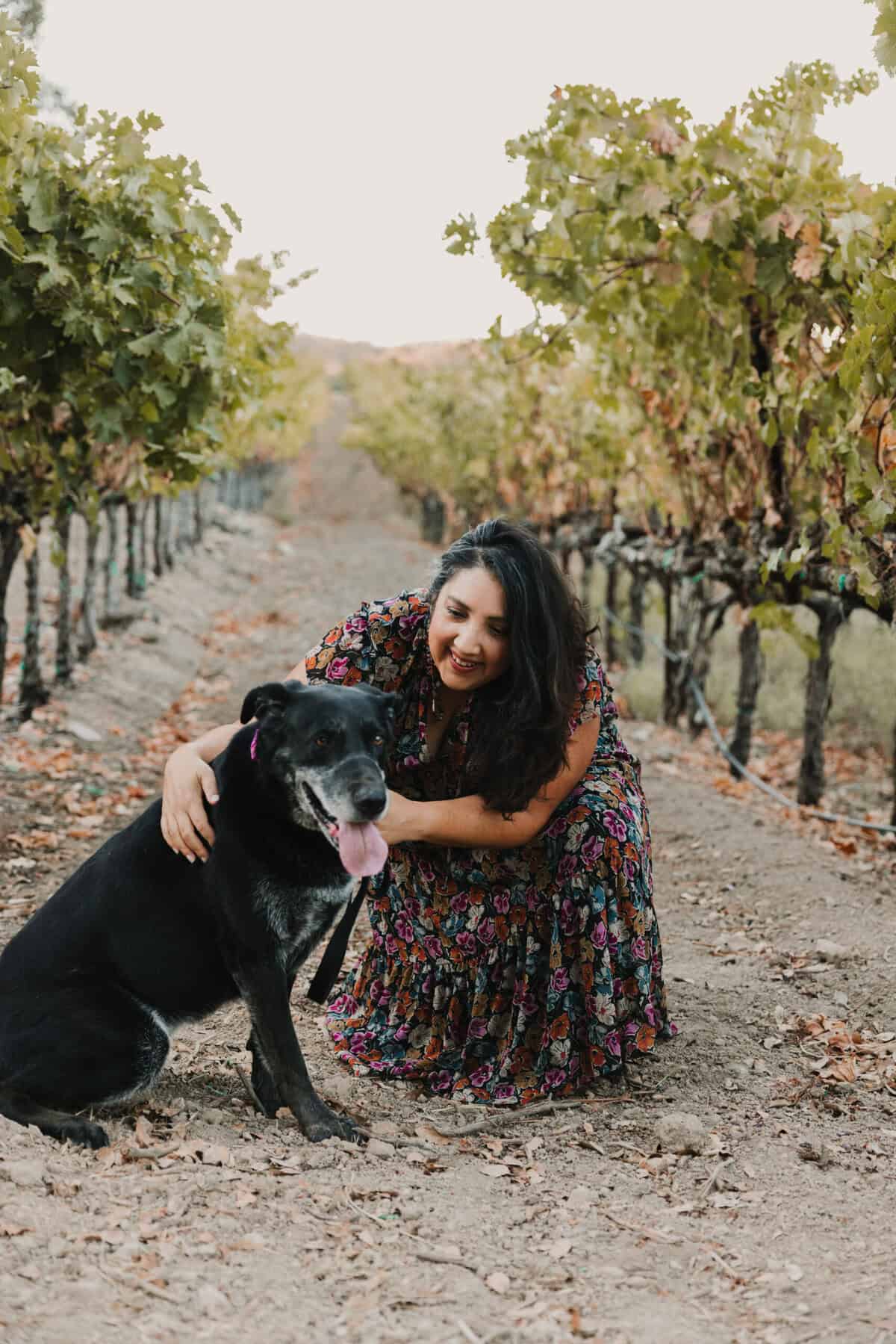 Photo of Frances in a floral dress kneeling beside her black labrador for an autumn photo against a backdrop of a vineyard with leaves of green and gold.